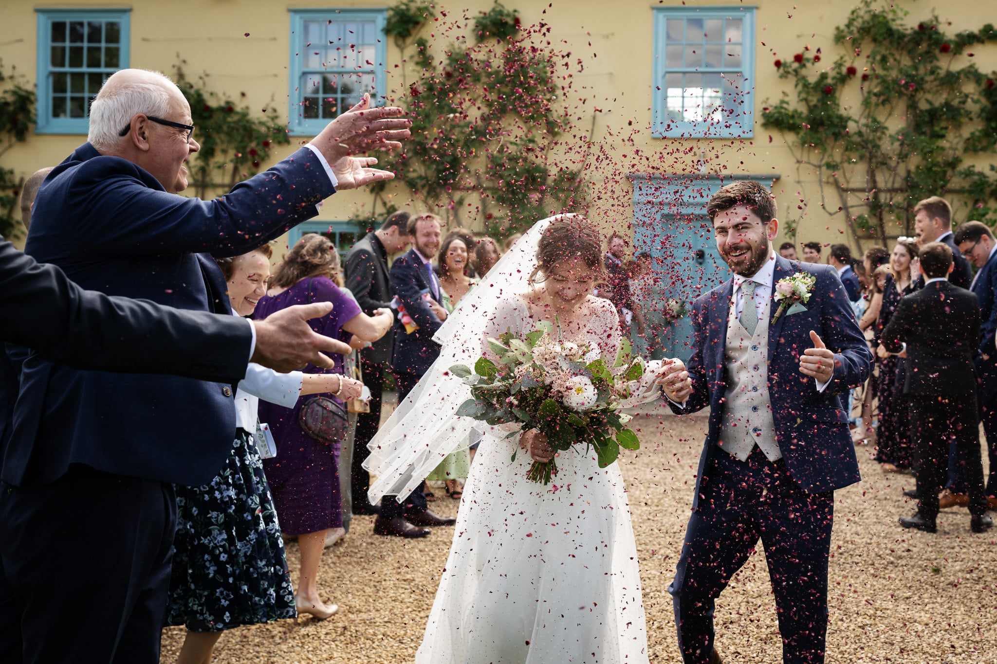 A bride and groom walk outside as guests throw confetti during a joyful South Farm wedding, with people smiling and a yellow building in the background.