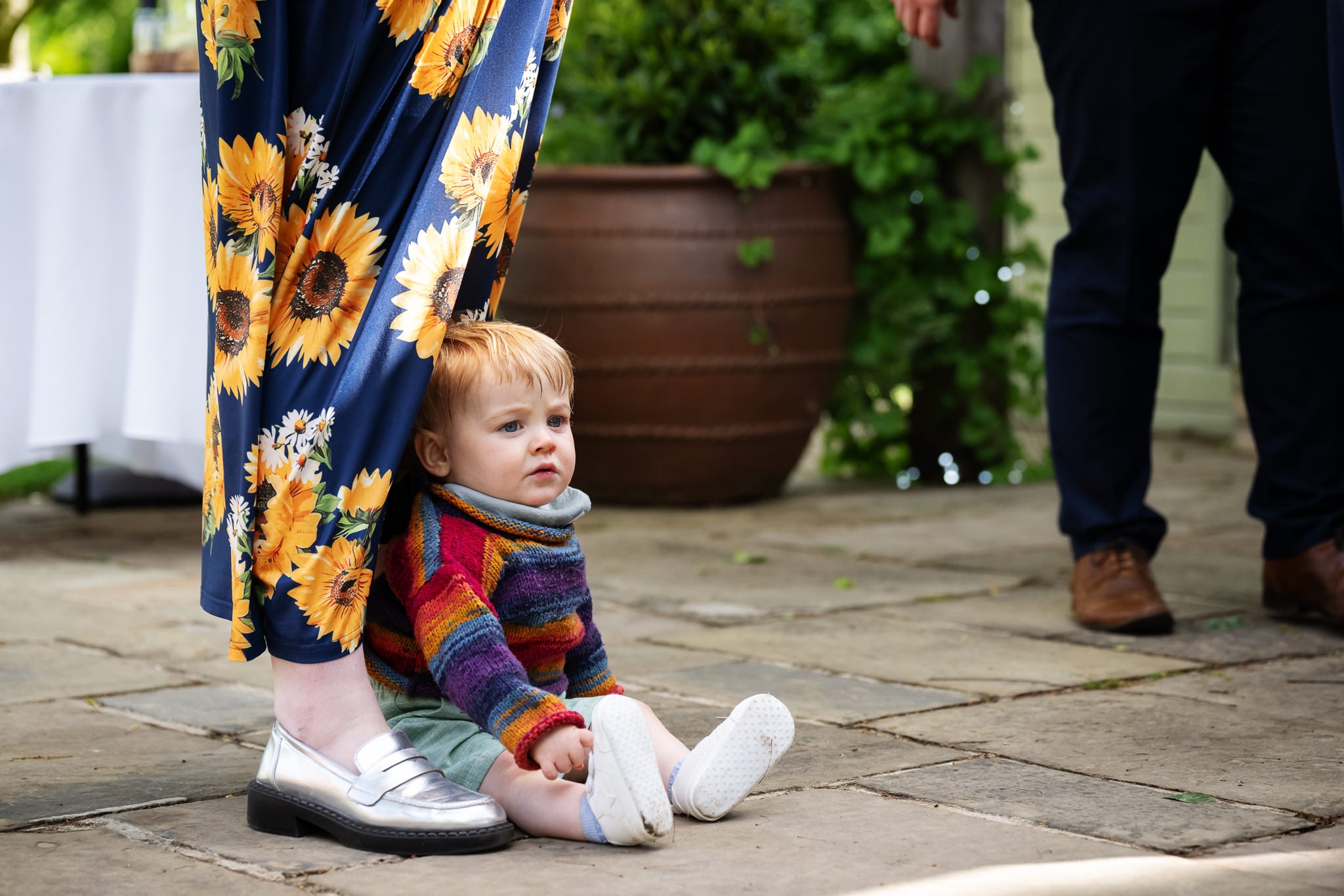 A young child in a colorful sweater sits on the ground at South Farm, leaning against an adult's legs dressed in sunflower-patterned pants and silver shoes.