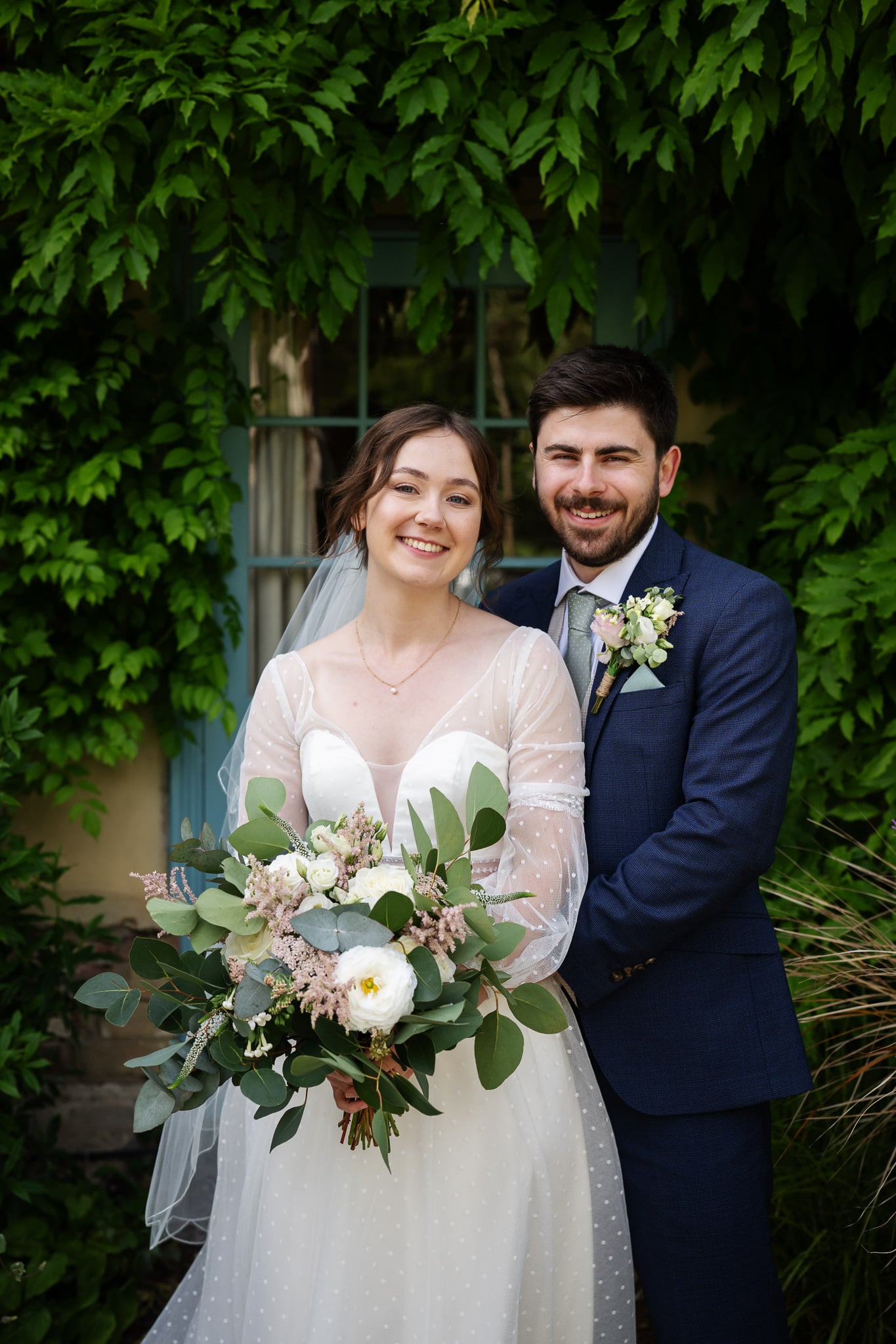 A bride in a white dress holds a bouquet and stands beside the groom in a navy suit, both smiling, in front of a window framed by green vines at South Farm.