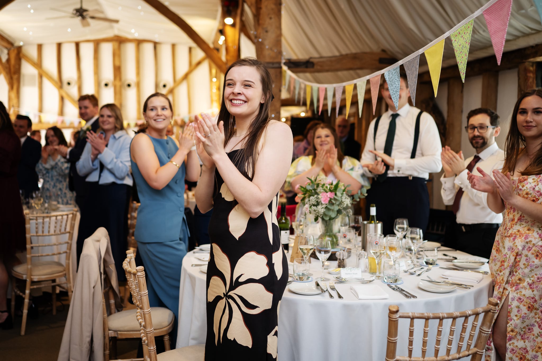 Guests in formal attire stand and applaud around decorated tables at South Farm, as natural light fills the room and bunting hangs overhead, celebrating the bride and groom at this elegant indoor event.
