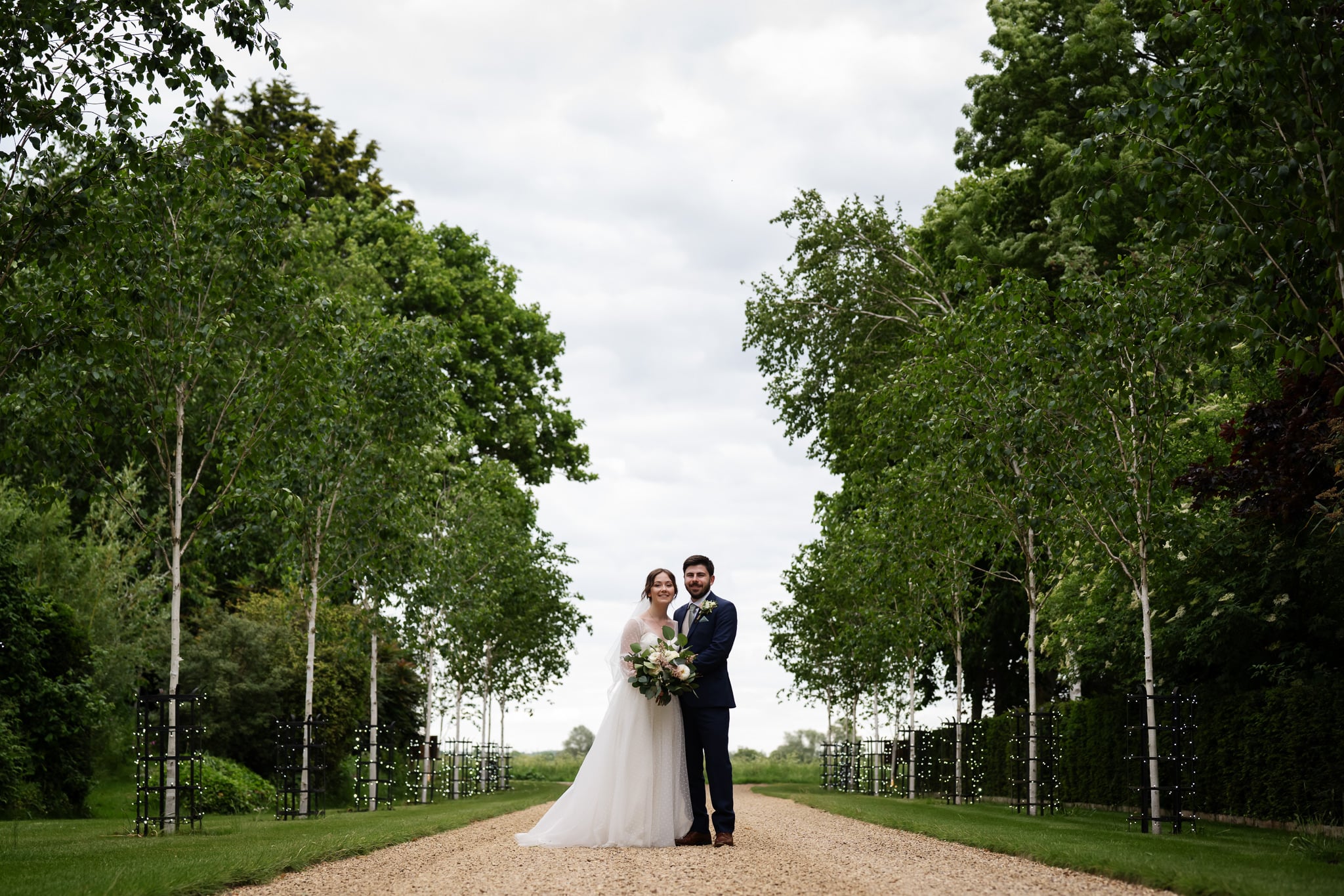 A bride and groom stand together on a gravel path lined with young trees, holding a bouquet, during their South Farm wedding in an outdoor setting filled with greenery and a cloudy sky.