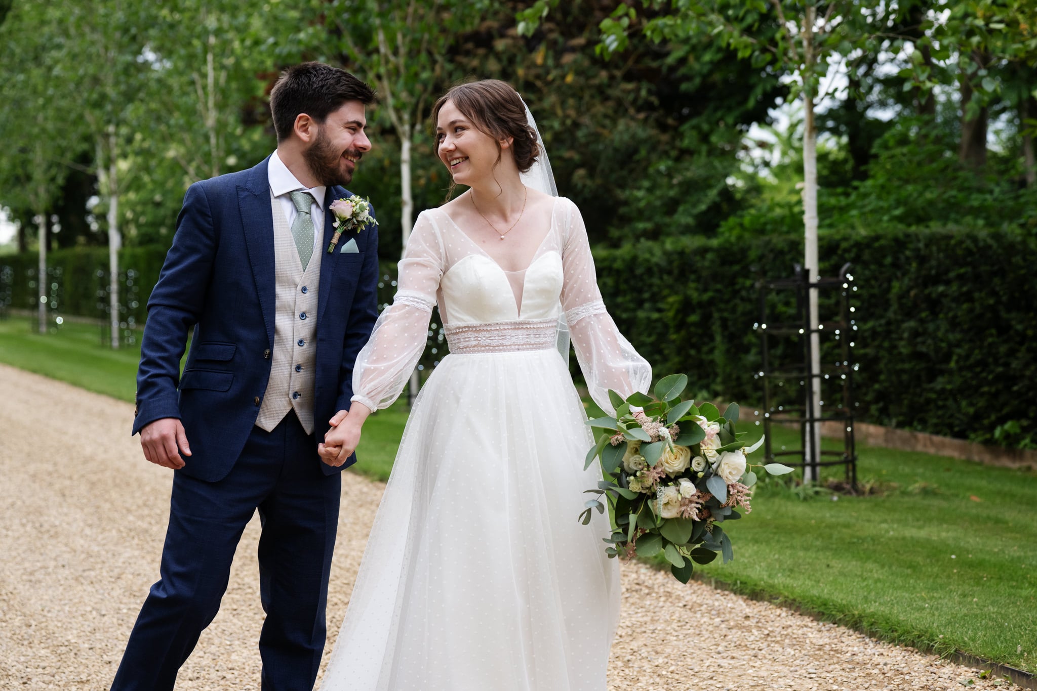 A bride in a white dress and a groom in a blue suit walk hand in hand on a gravel path, surrounded by greenery at their beautiful South Farm wedding.