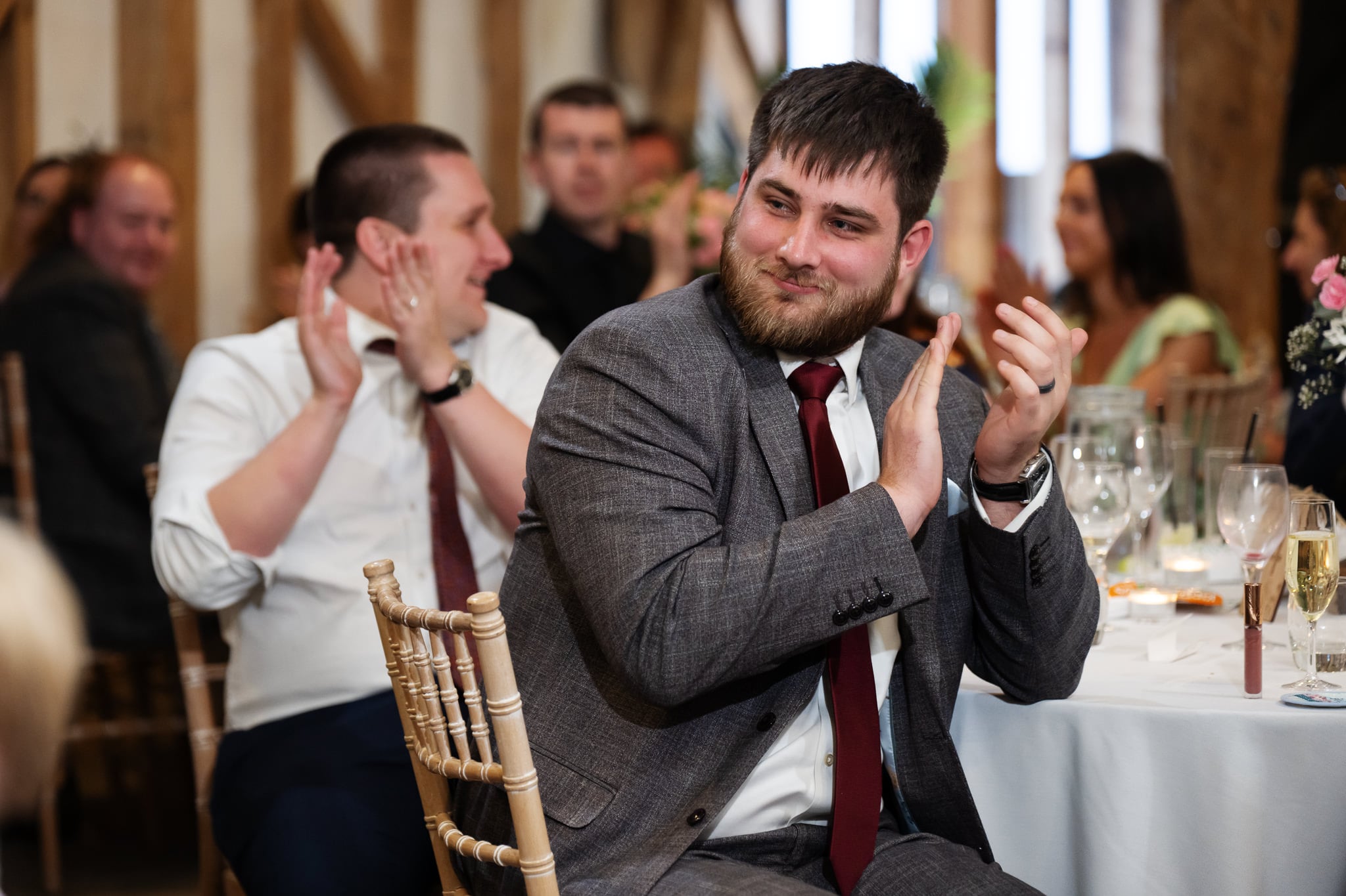 A man in a gray suit and red tie, possibly the groom, sits at a round table clapping, with other people—perhaps guests at a South Farm wedding—seated and applauding in the background.
