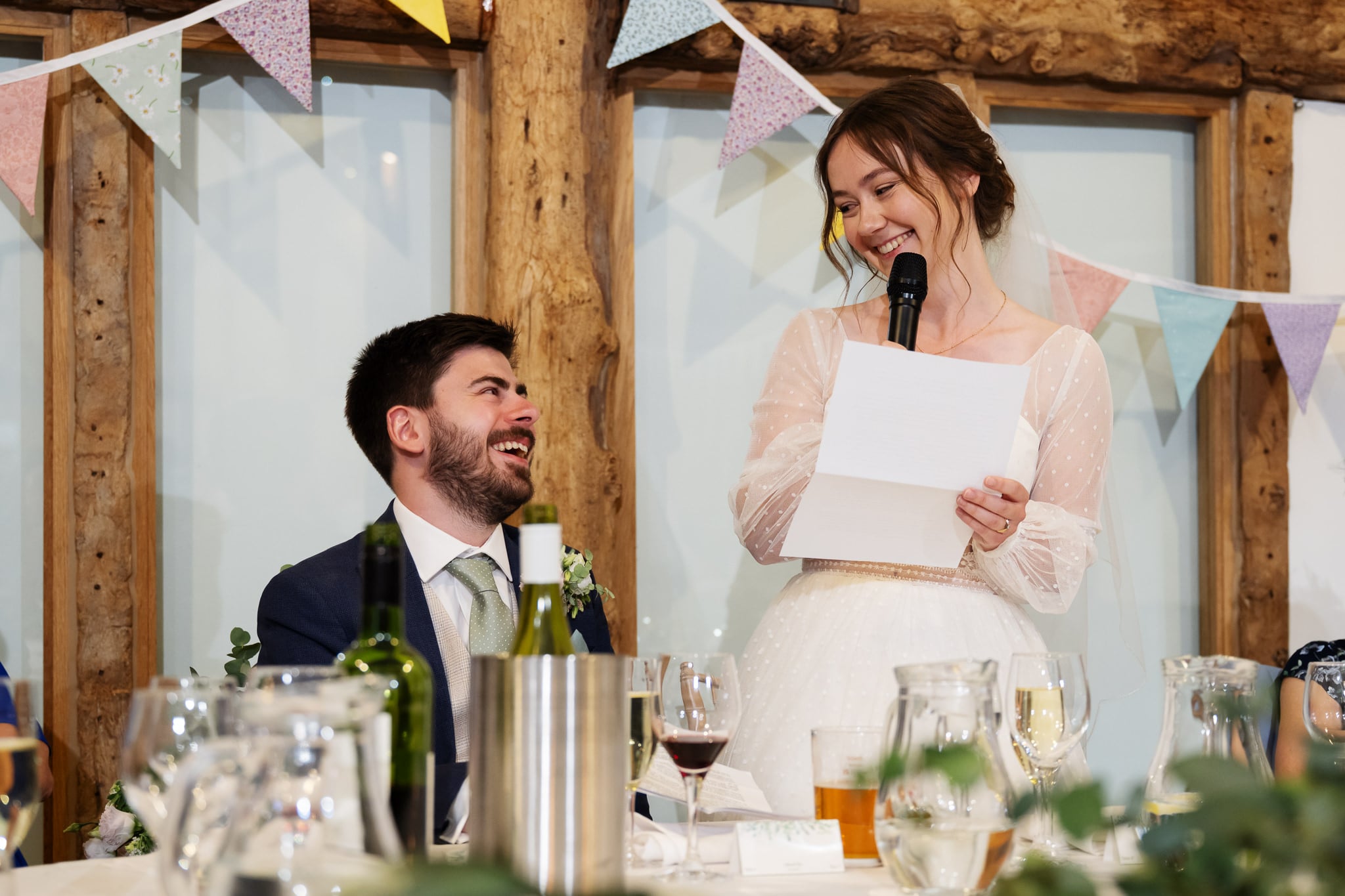 At a South Farm wedding, the Bride in a white dress reads from a paper with a microphone while a man in a suit sits beside her, smiling; drinks and decorations are on the table in front of them.