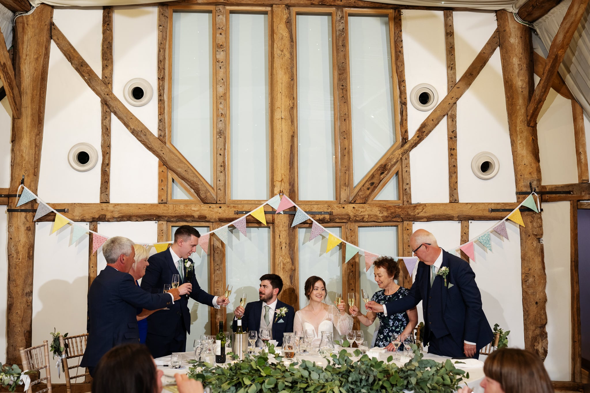 A group of people, including the Bride and Groom, sit at a table in front of tall windows and wooden beams, raising glasses for a toast at their joyful South Farm wedding reception.