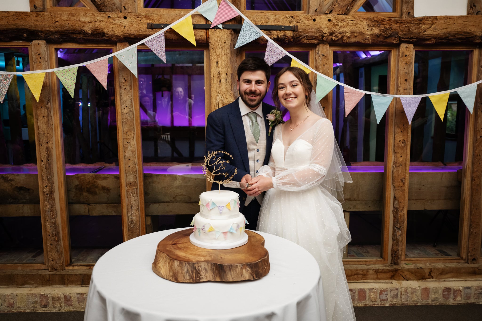 The bride and groom stand together, smiling as they cut a three-tiered wedding cake on a wooden table at South Farm, with bunting and rustic wooden beams in the background.