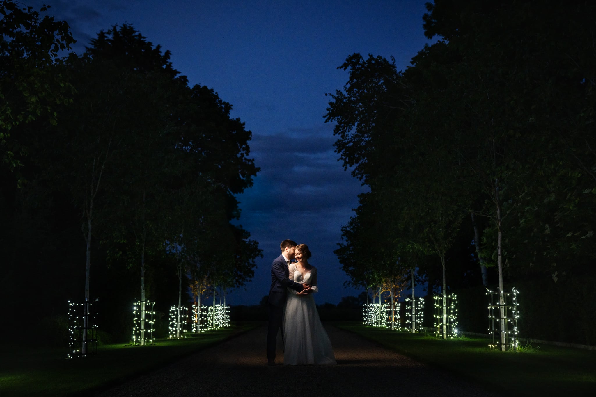 A Bride and Groom in wedding attire embrace on a tree-lined path at South Farm at night, illuminated by small white lights wrapped around the trunks.