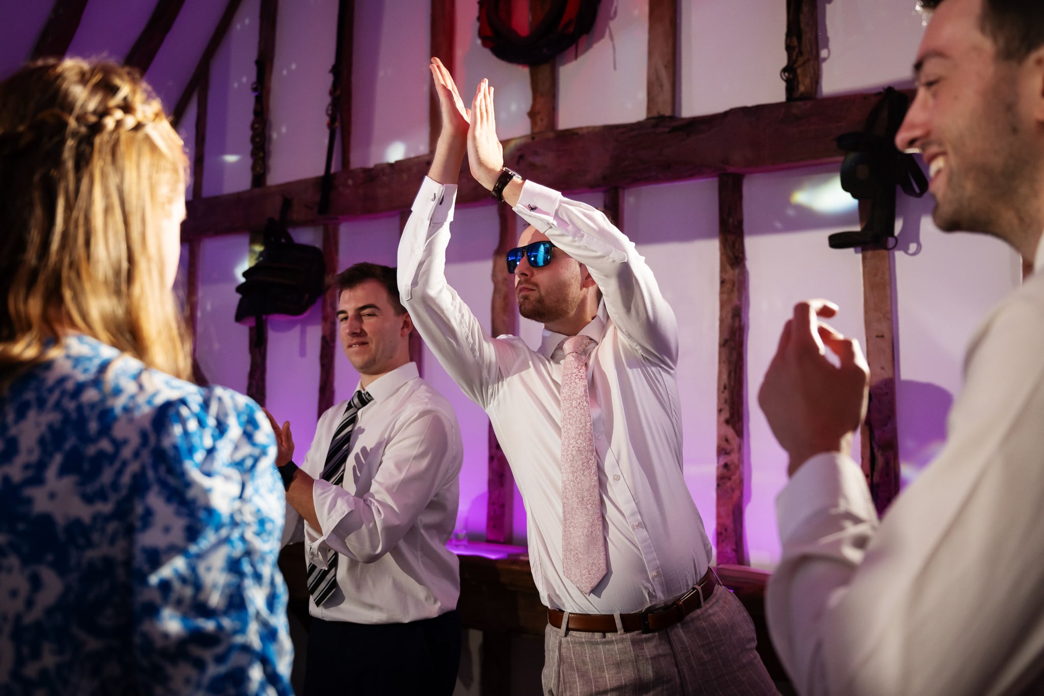 A man in sunglasses, possibly the groom, claps above his head while others stand nearby, smiling and dressed in formal attire at an indoor South Farm event.