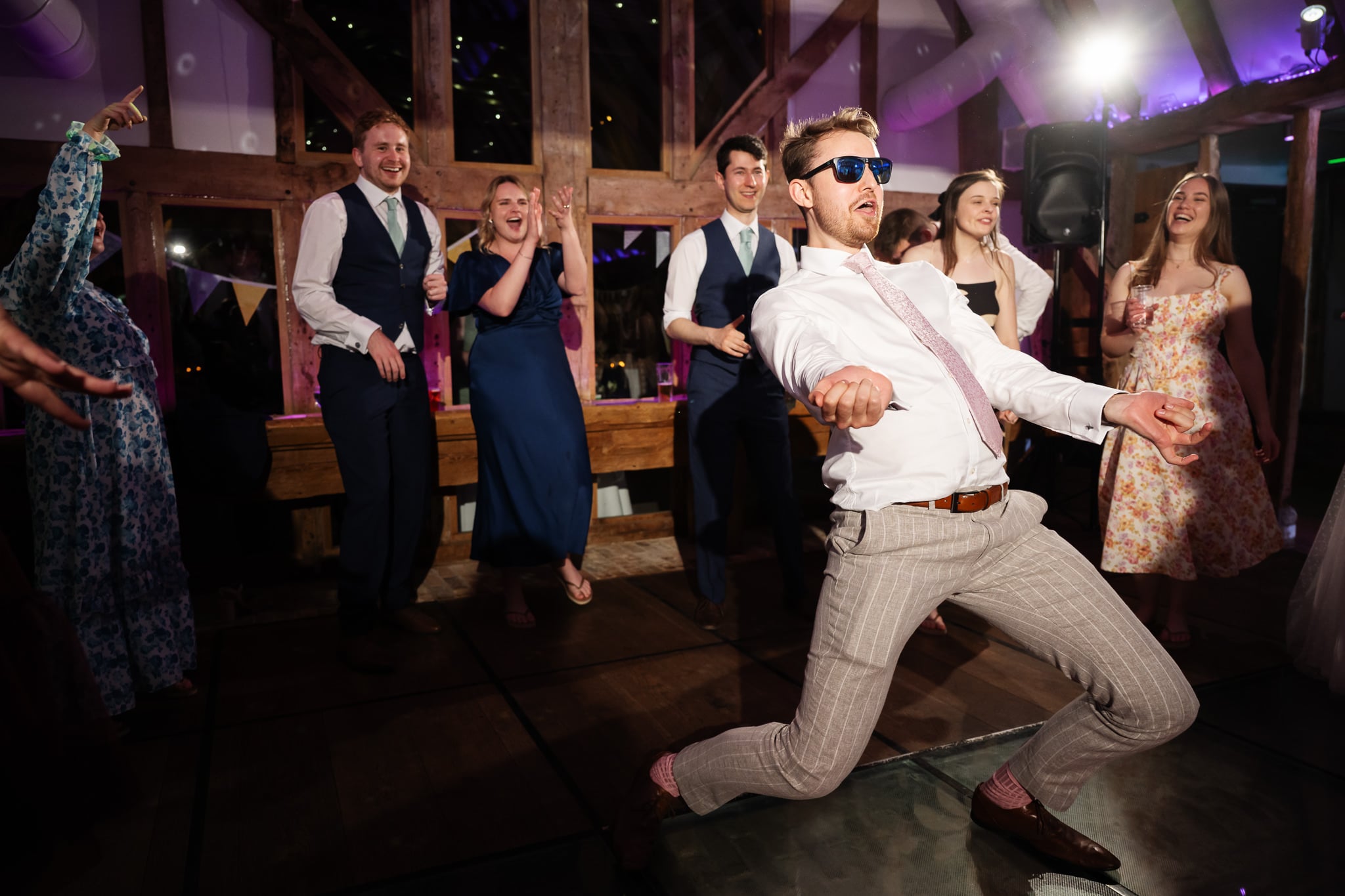 A groom wearing sunglasses dances energetically in front of a group of people dressed in formal attire at an indoor South Farm event.