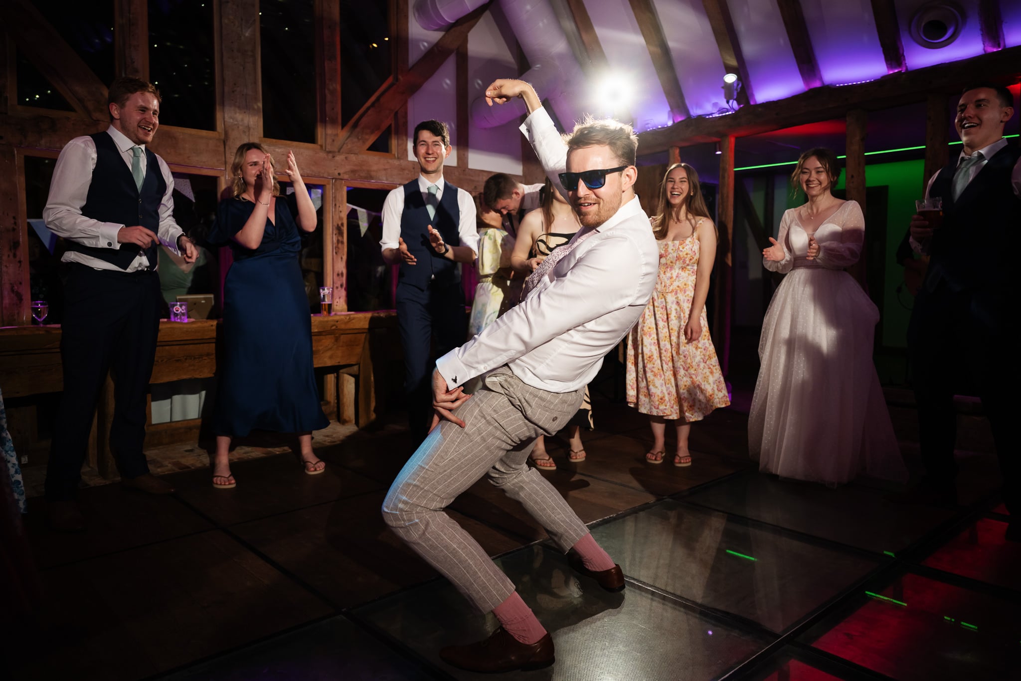 A man in sunglasses strikes a playful dance pose on a lit dance floor at a South Farm wedding, surrounded by smiling guests and the bride, as everyone claps at this festive indoor event.