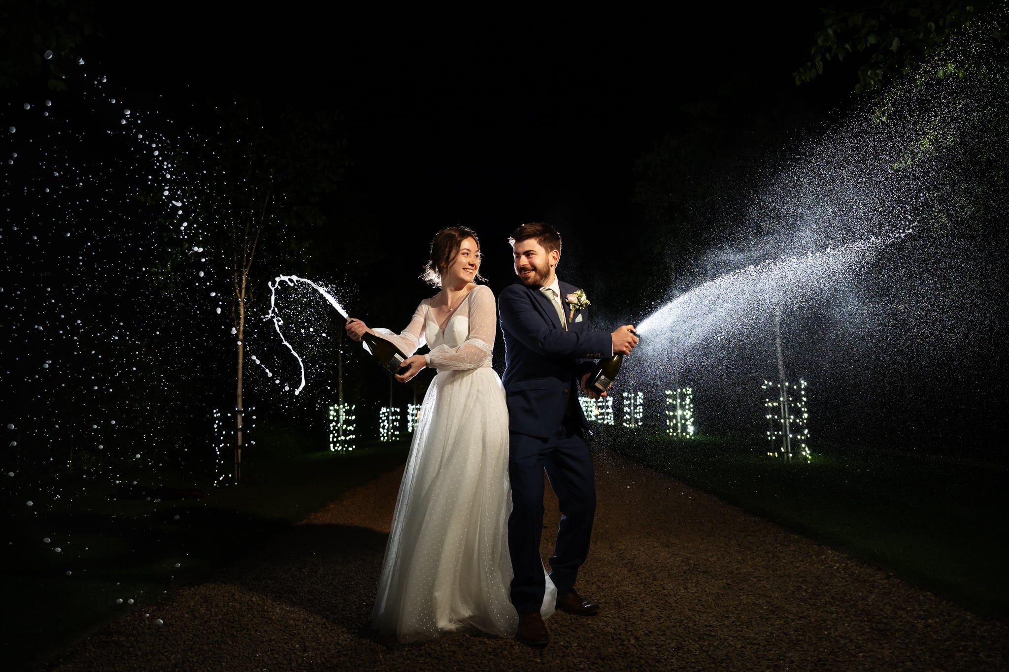 A bride and groom in formal attire spray champagne bottles outdoors at night at South Farm, with illuminated trees in the background.