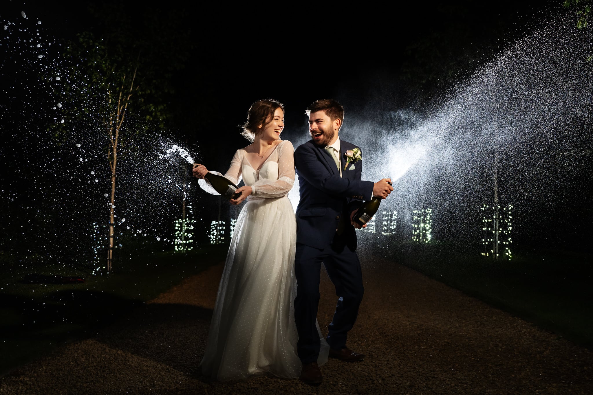 A bride and groom in formal attire spray champagne outdoors at night during their South Farm wedding, both smiling and surrounded by illuminated trees.