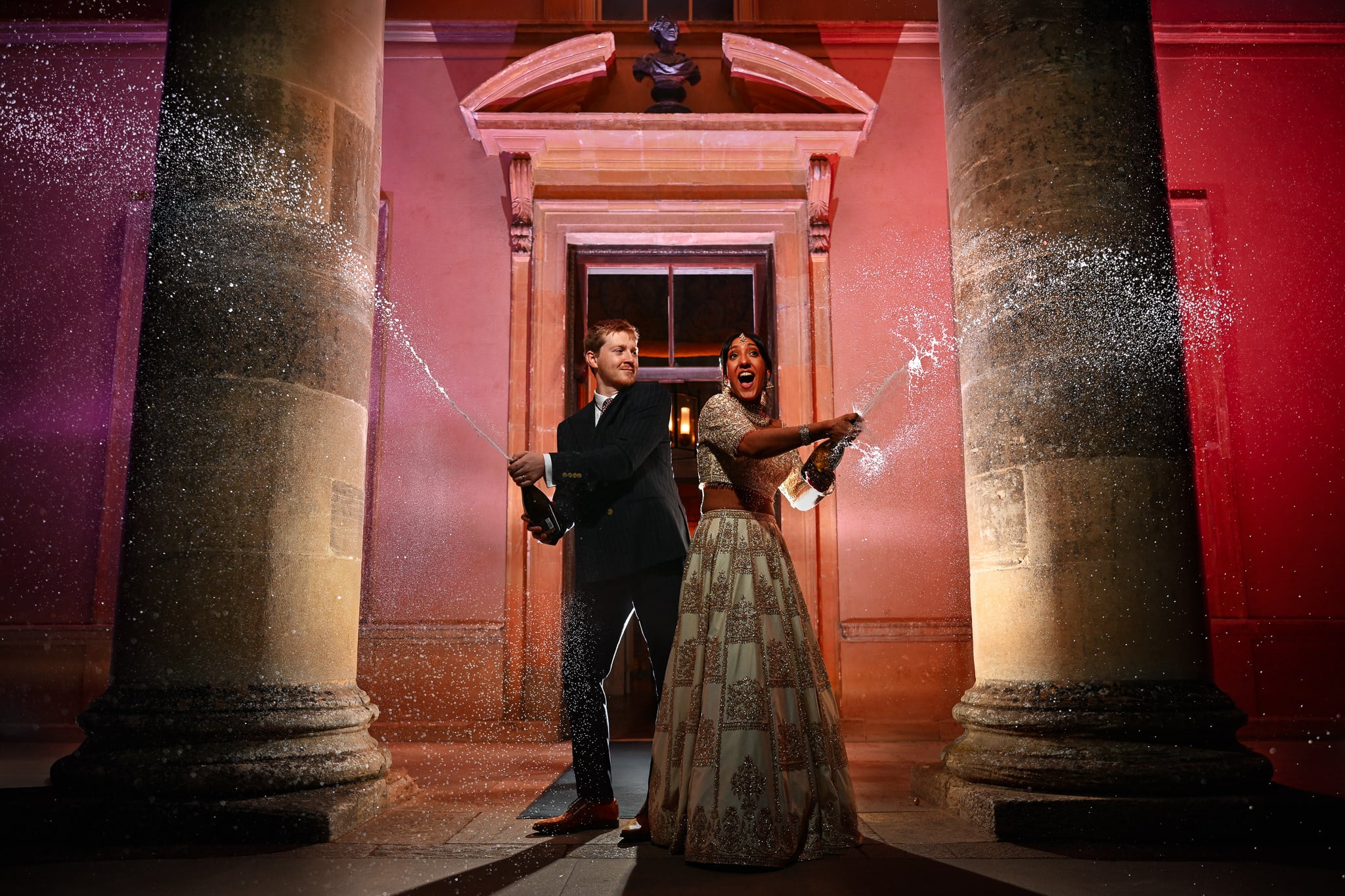 A portrait of the bride and groom stood on the steps of Stowe House whilst spraying champagne