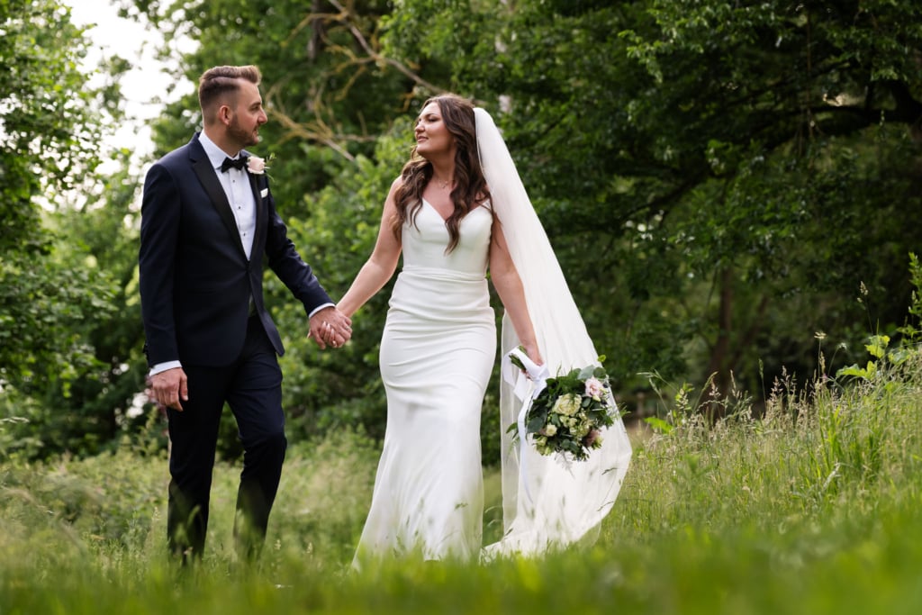 A bride in a white dress and veil holds a bouquet and walks hand in hand with a groom in a black suit through the lush, grassy grounds of their beautiful Tewinbury wedding in Hertfordshire.