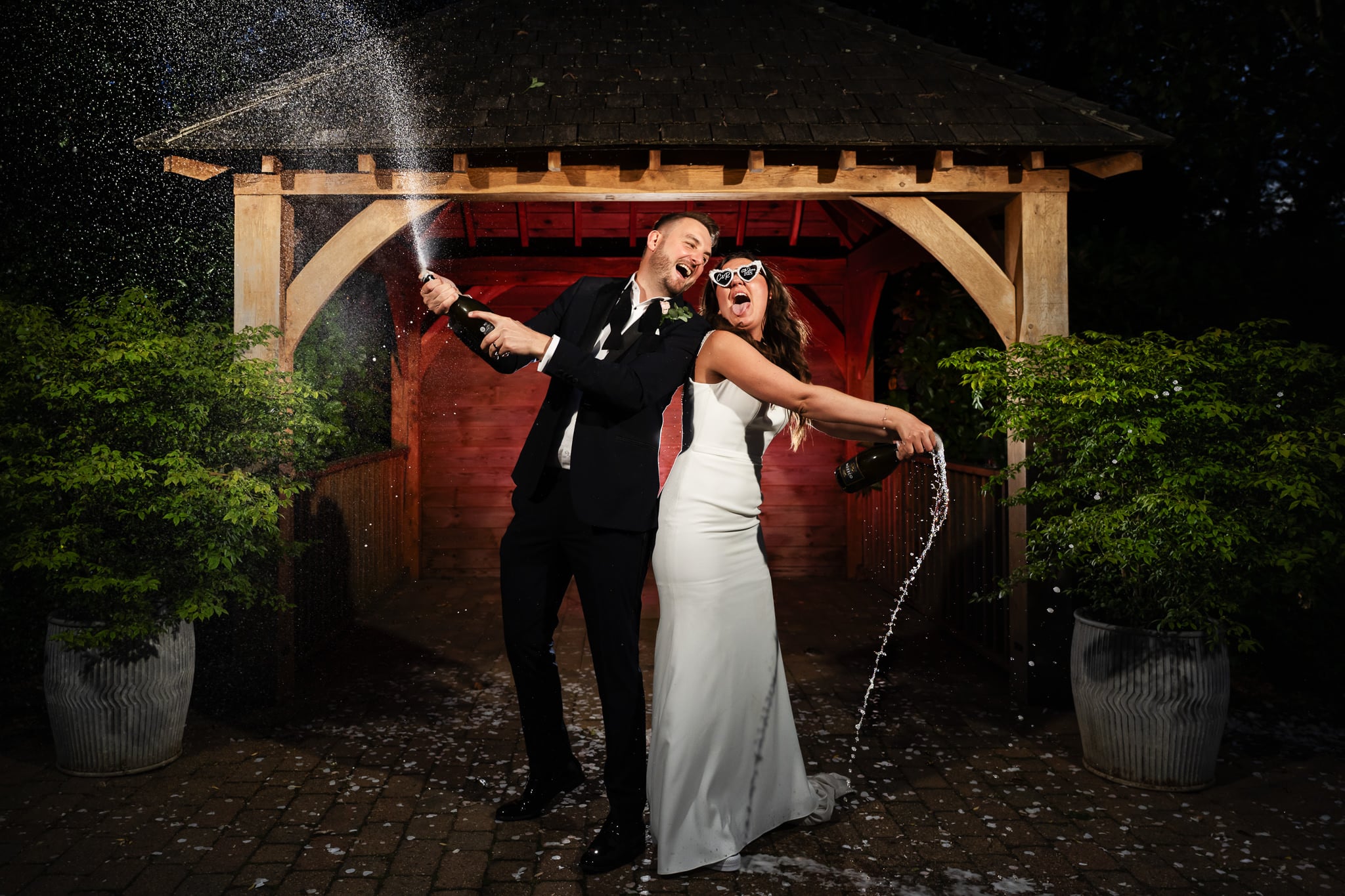 A bride and groom in formal attire celebrate outdoors at their Tewinbury wedding, spraying champagne and smiling together under a wooden gazebo.