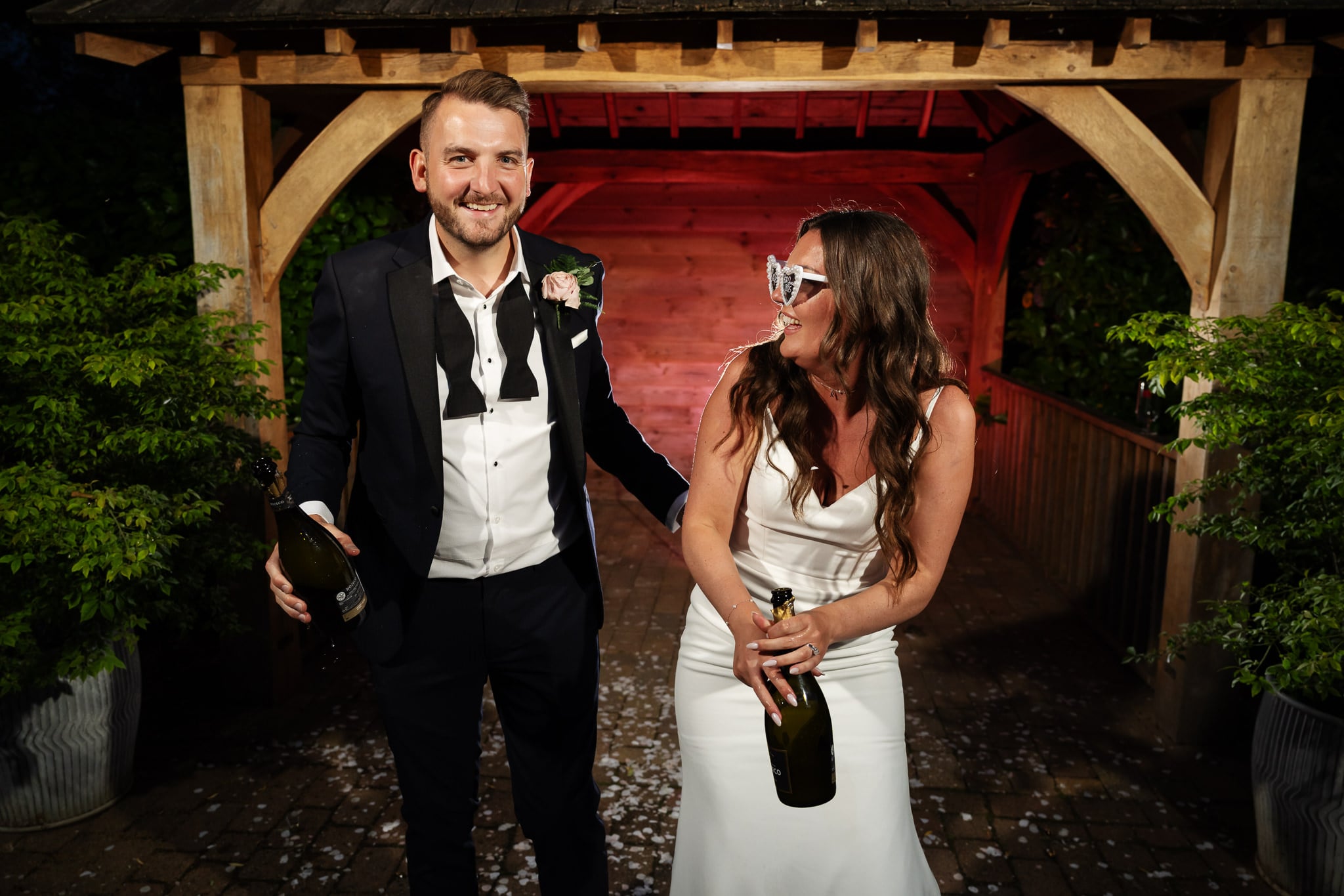 A man in a suit and the smiling Bride in a white dress hold champagne bottles under a wooden gazebo at their barn venue, with scattered petals on the ground.