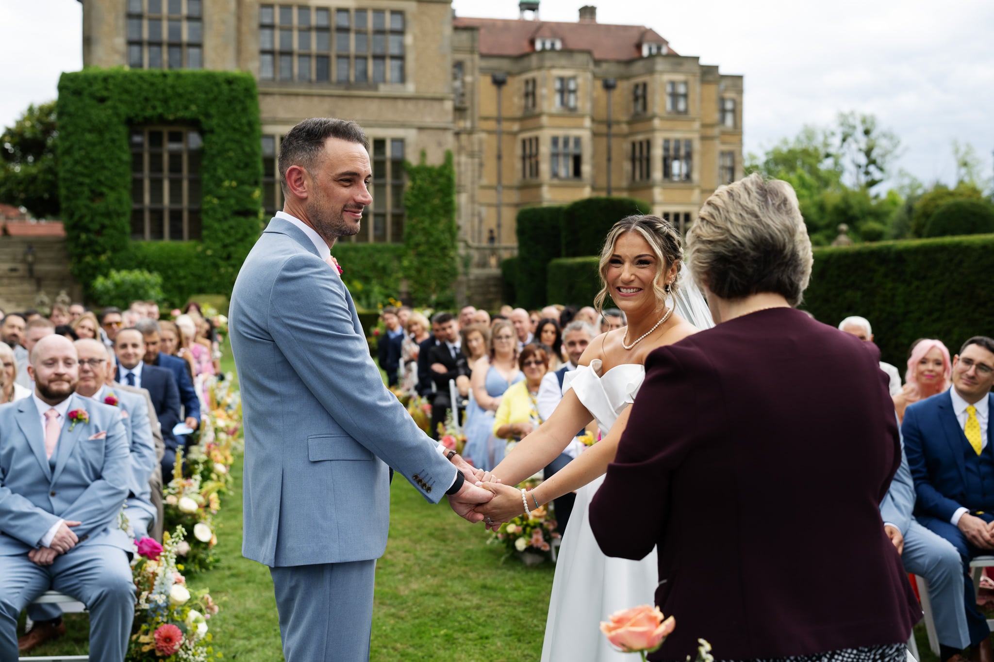Bride and groom holding hands and smiling at their outdoor Fanhams Hall wedding, with guests seated nearby.