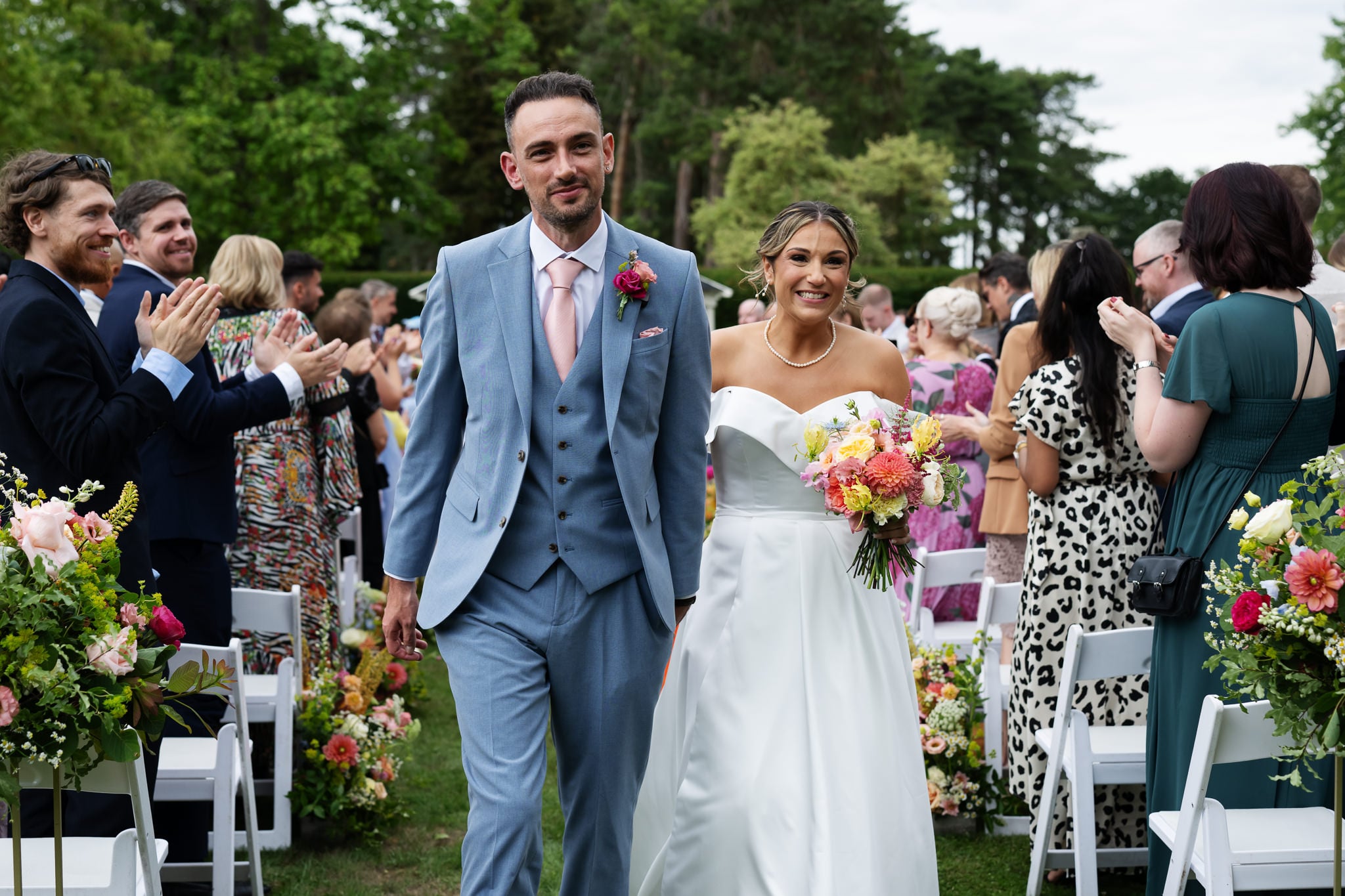Smiling bride and groom walk down the aisle, surrounded by happy guests and colourful flowers.