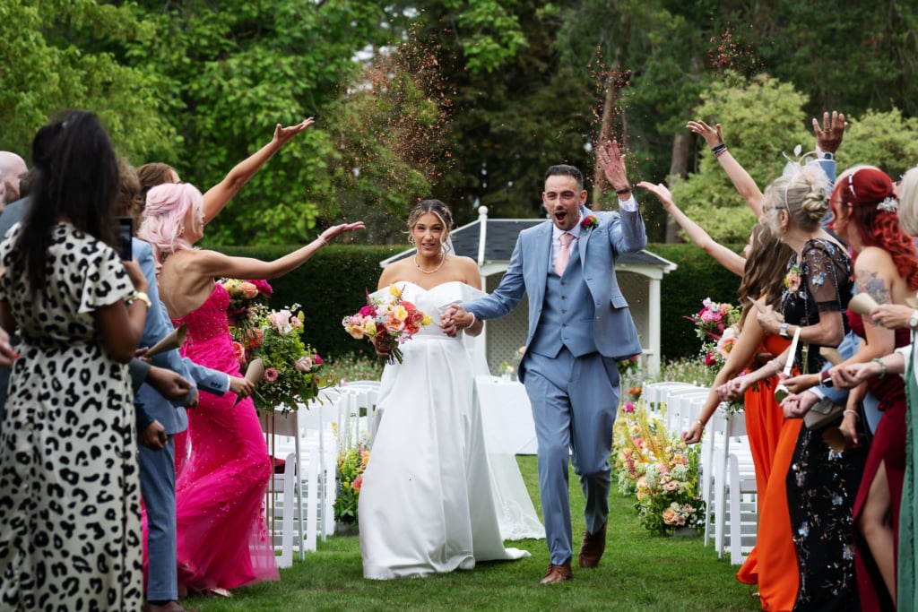 Bride and groom walk down the Fanhams Hall aisle as guests throw confetti outdoors.