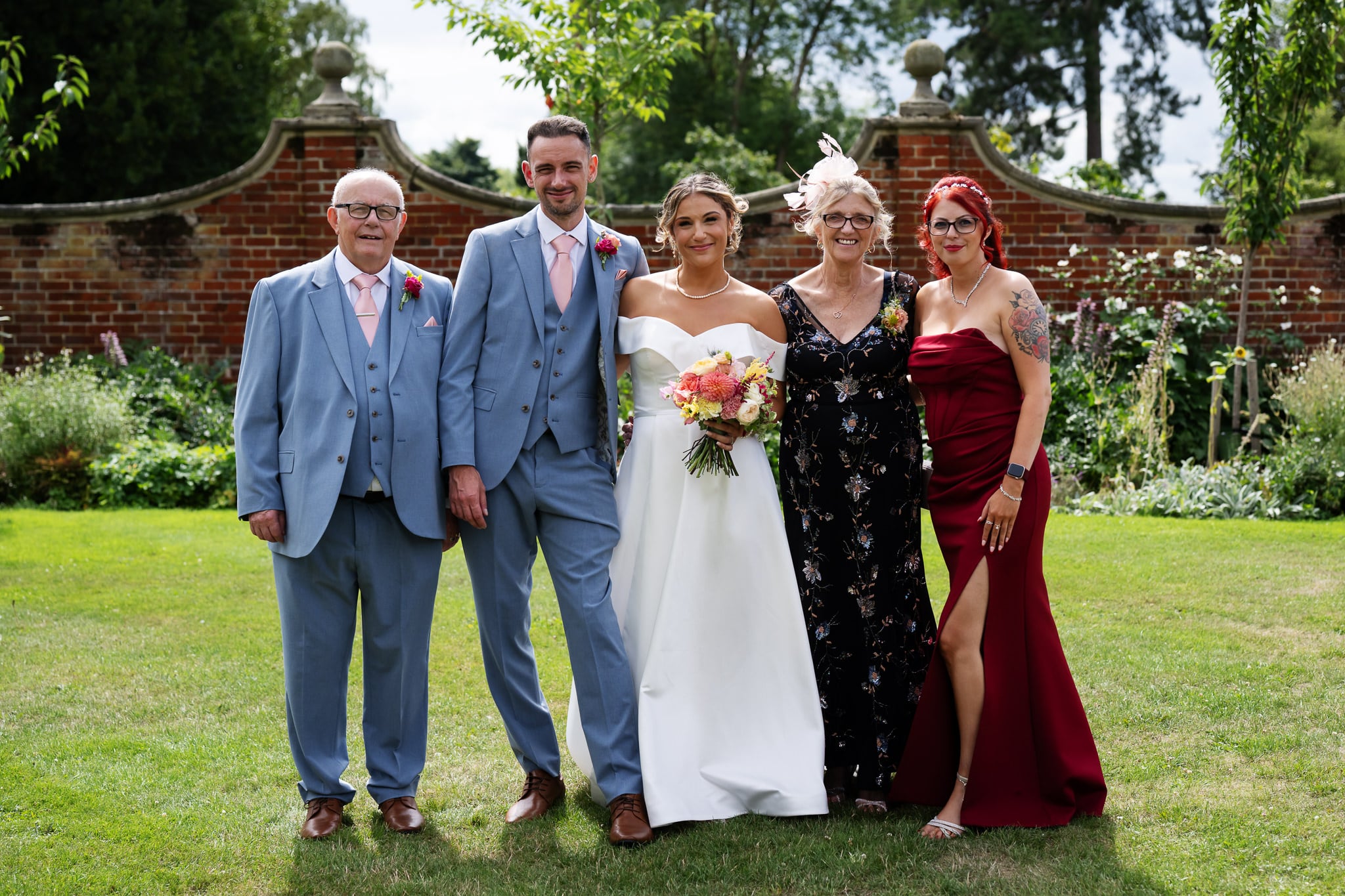 Five formally dressed people, including bride and groom, pose outdoors at a wedding with greenery and a brick wall behind them.