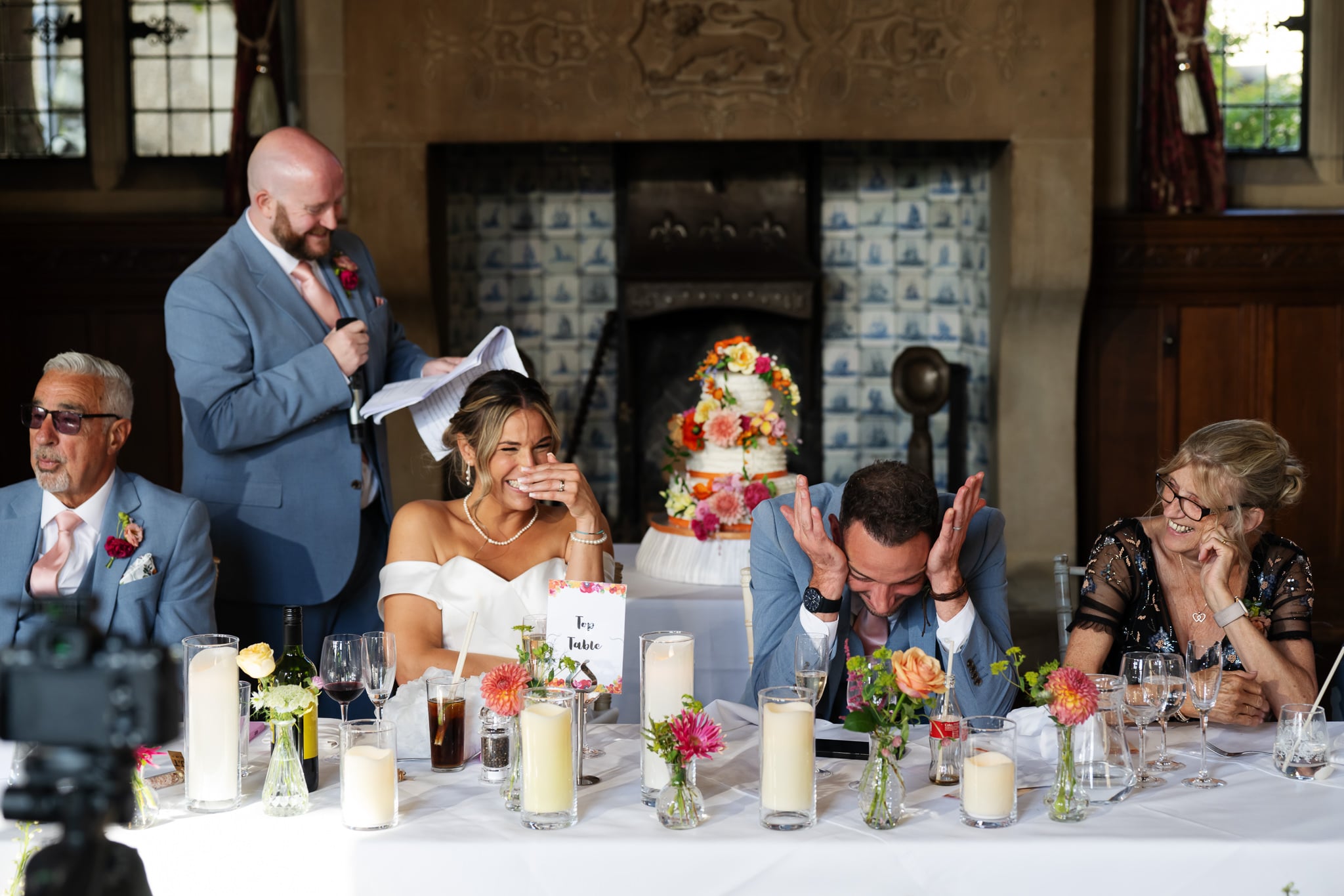 At Fanhams Hall, a wedding speech makes the bride laugh and the groom cover his face, amusing guests at the reception table.