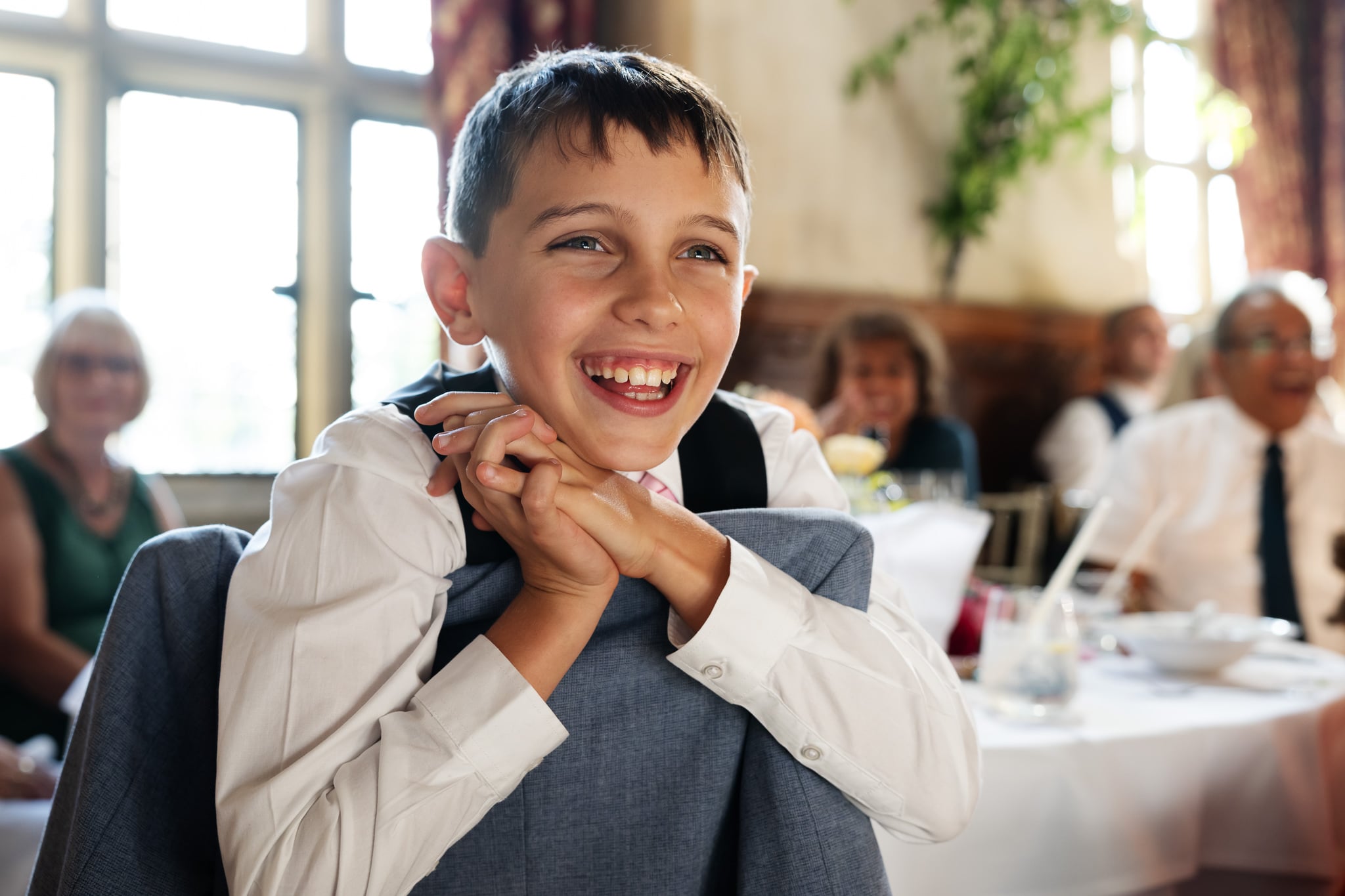 Smiling boy in formal wear leans on a chair at an indoor wedding with guests in the background at Fanhams Hall.