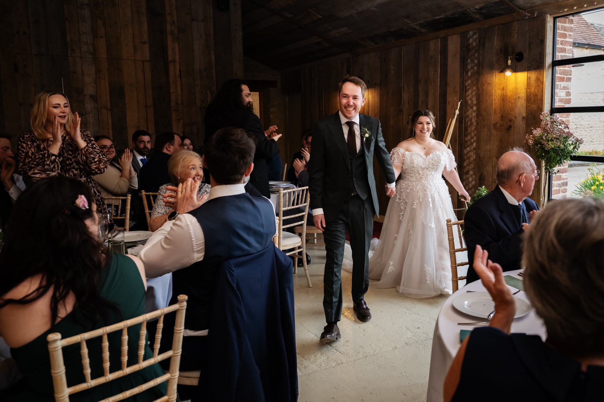 A bride and groom enter the rustic Stratton Court Barn wedding reception room, smiling and holding hands, as seated guests applaud and watch them.