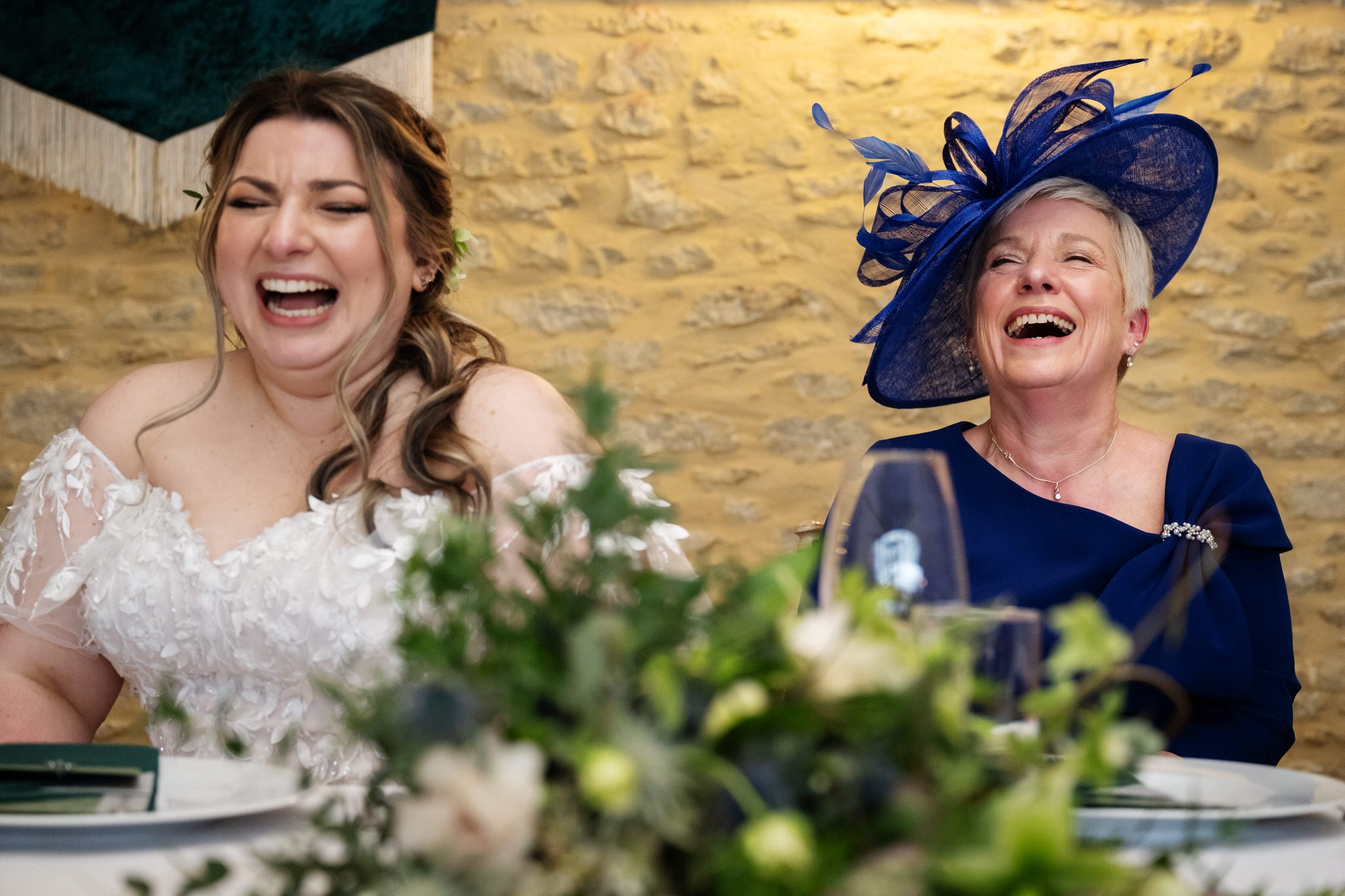 Two women, one in a white wedding dress and one in a blue dress with a matching hat, sit at a table laughing at Stratton Court Barn. A floral centerpiece is visible in the foreground, adding to the joyful wedding atmosphere.
