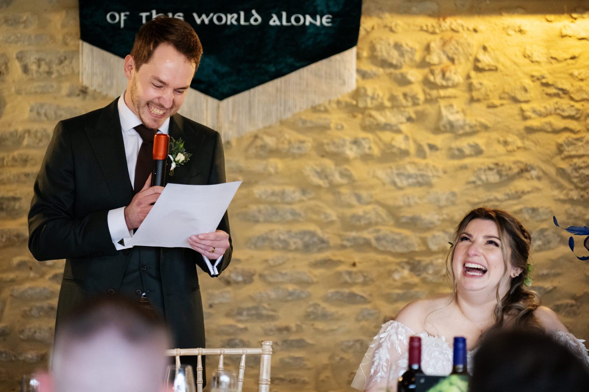 At Stratton Court Barn, the groom in a suit reads from a paper into a microphone as the bride in a white dress sits and laughs, with a stone wall and a banner in the background.
