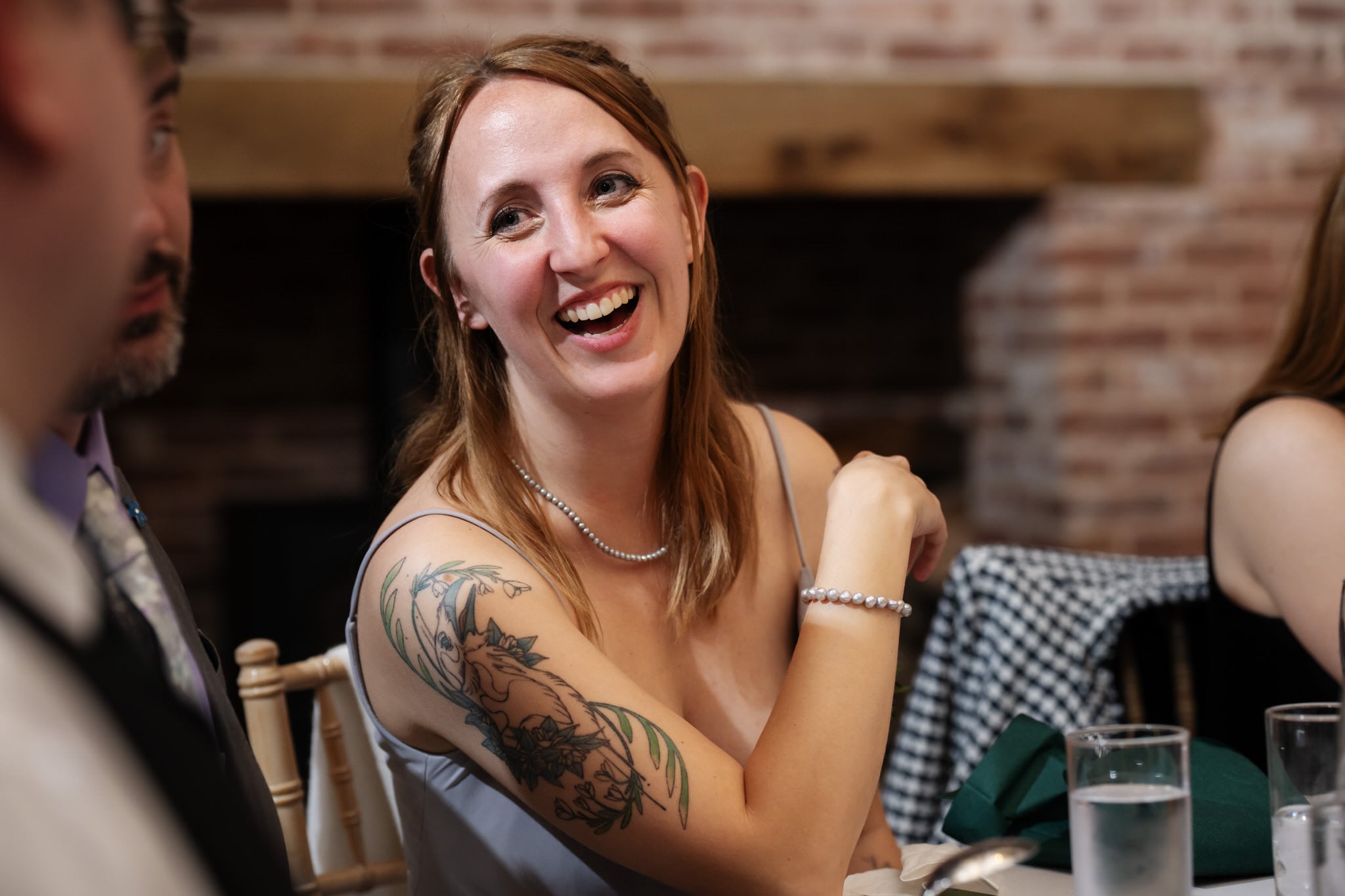 A woman with a floral tattoo on her arm smiles and chats with others at a table set with glasses of water and silverware during a wedding at Stratton Court Barn.