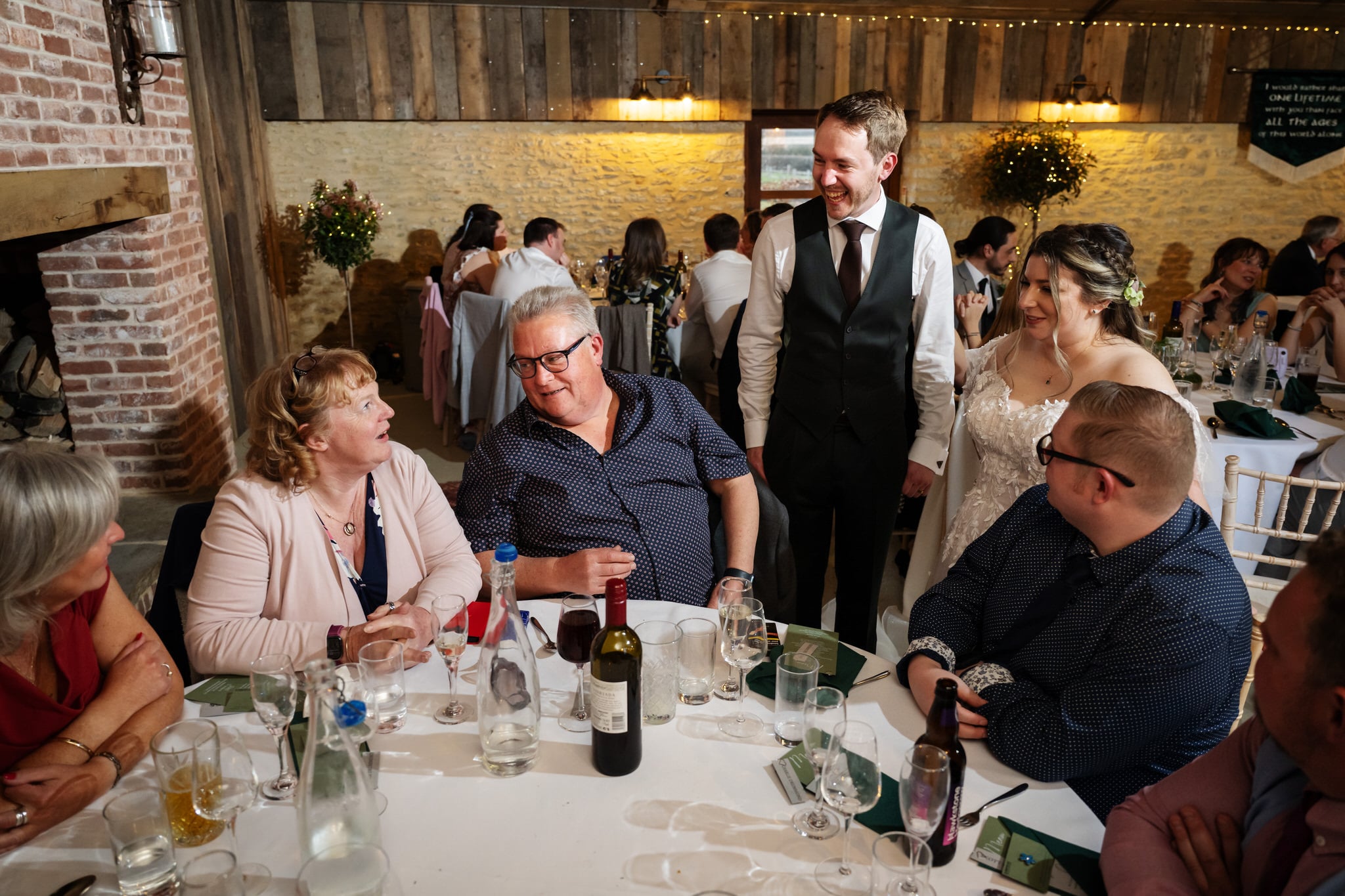 A bride and groom stand by a table, smiling and talking with seated wedding guests in the warmly lit reception hall at Stratton Court Barn.