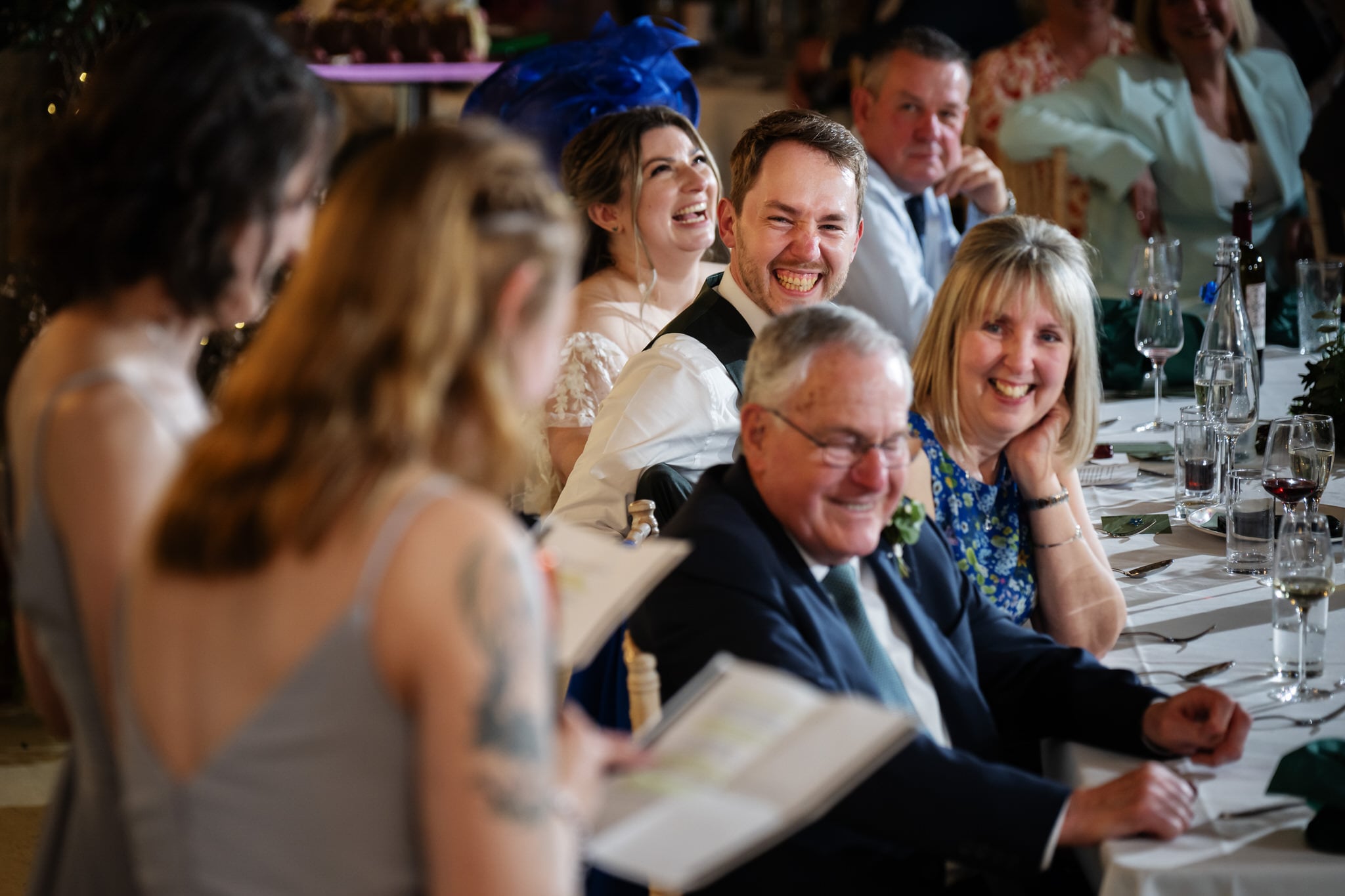 A joyful wedding reception at Stratton Court Barn, where guests seated at a table smile and laugh as someone reads heartfelt notes to the bride and groom.