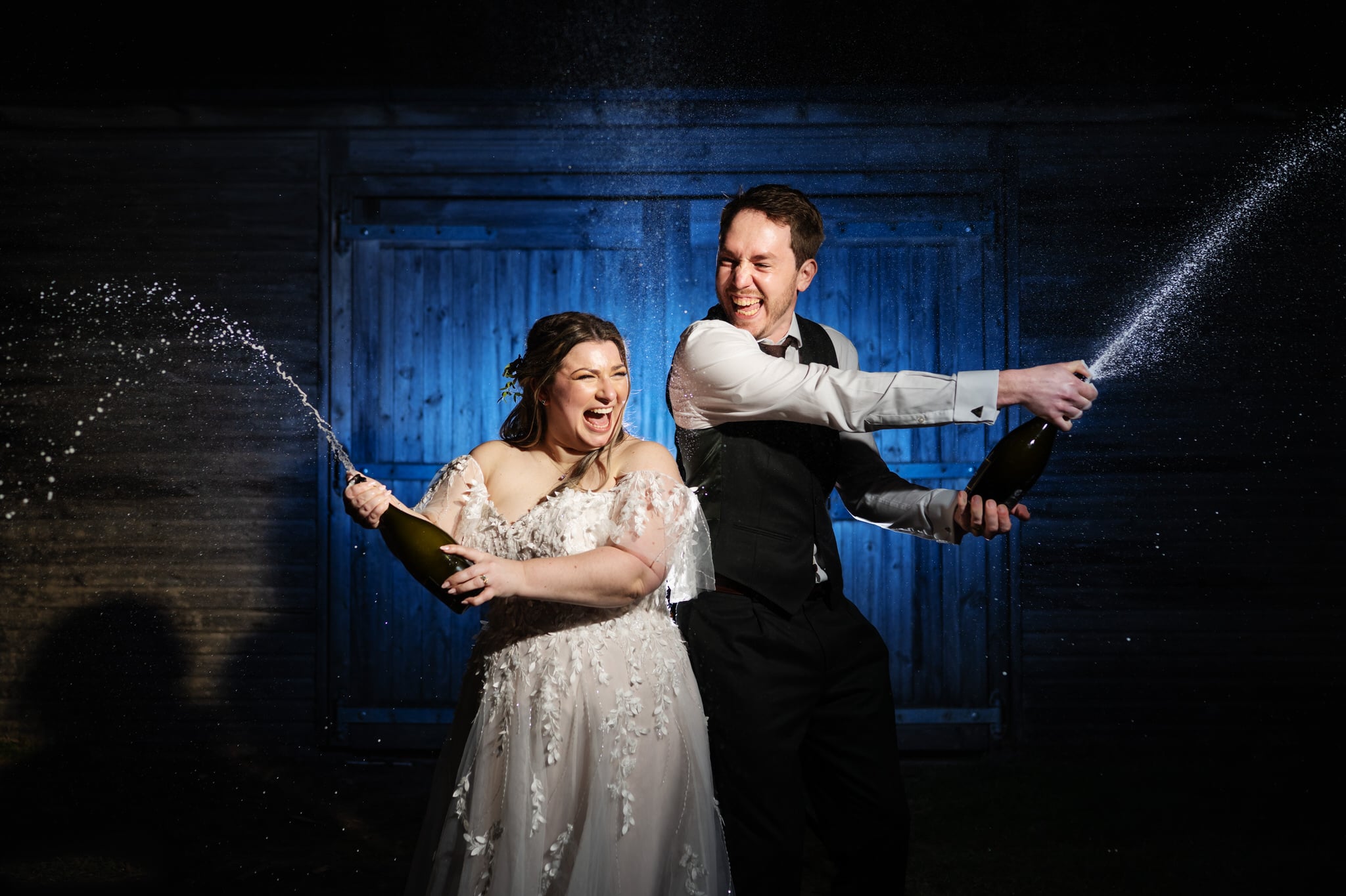 A bride and groom stand outside at night at Stratton Court Barn, smiling and celebrating as they spray champagne in front of a blue-lit wooden wall.