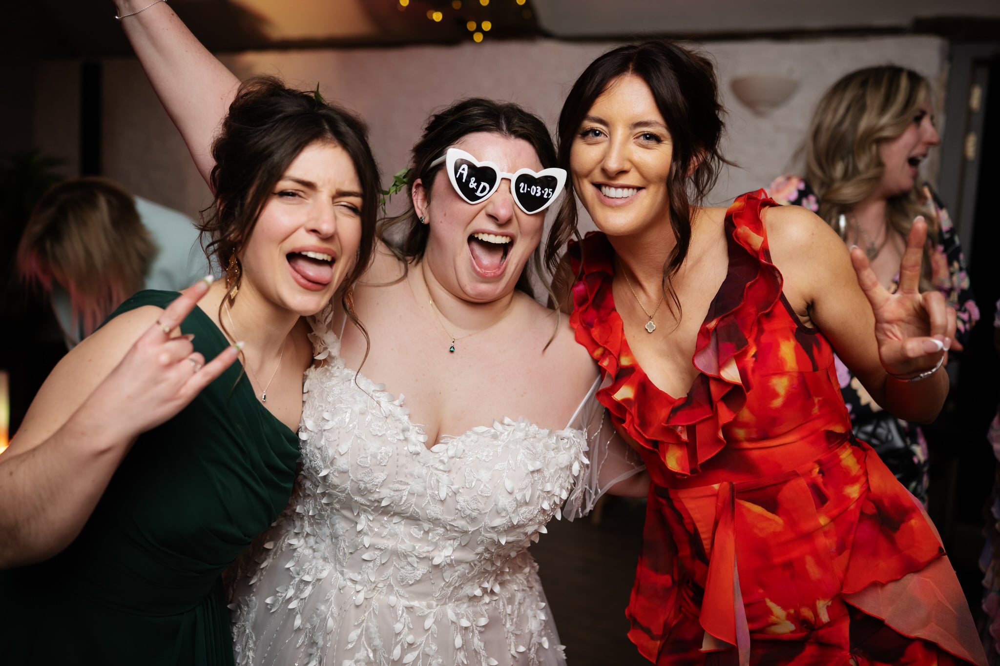 Three women pose and smile at a wedding party; one in a white dress—likely the bride—wears heart-shaped sunglasses reading "AED 8-3-23" and flashes a peace sign, celebrating at Stratton Court Barn.