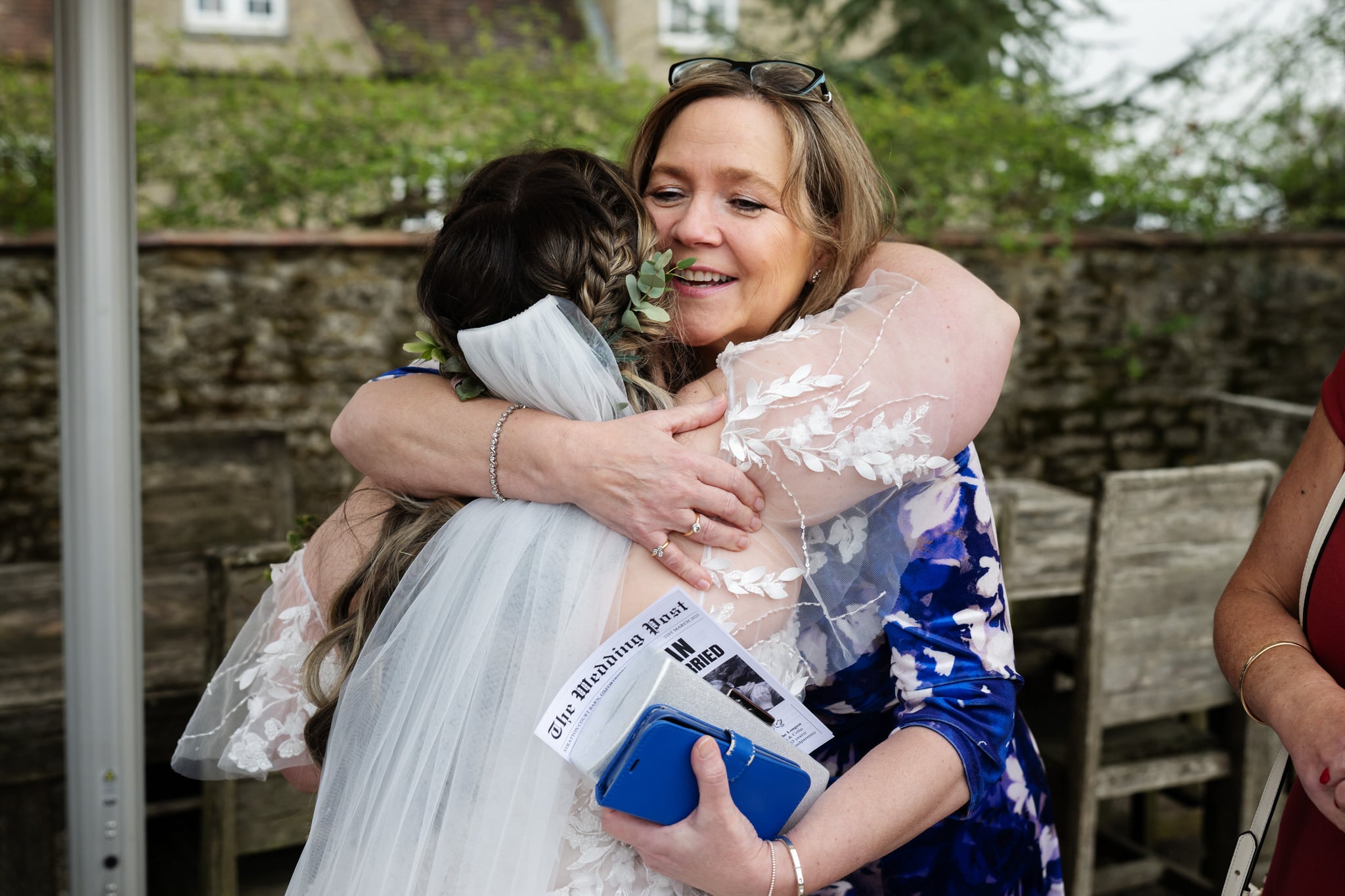 Two women embrace outdoors; one is the bride in a wedding veil holding "The Wedding Post," while the other smiles in a blue floral dress, holding a phone and glasses—captured at Stratton Court Barn.