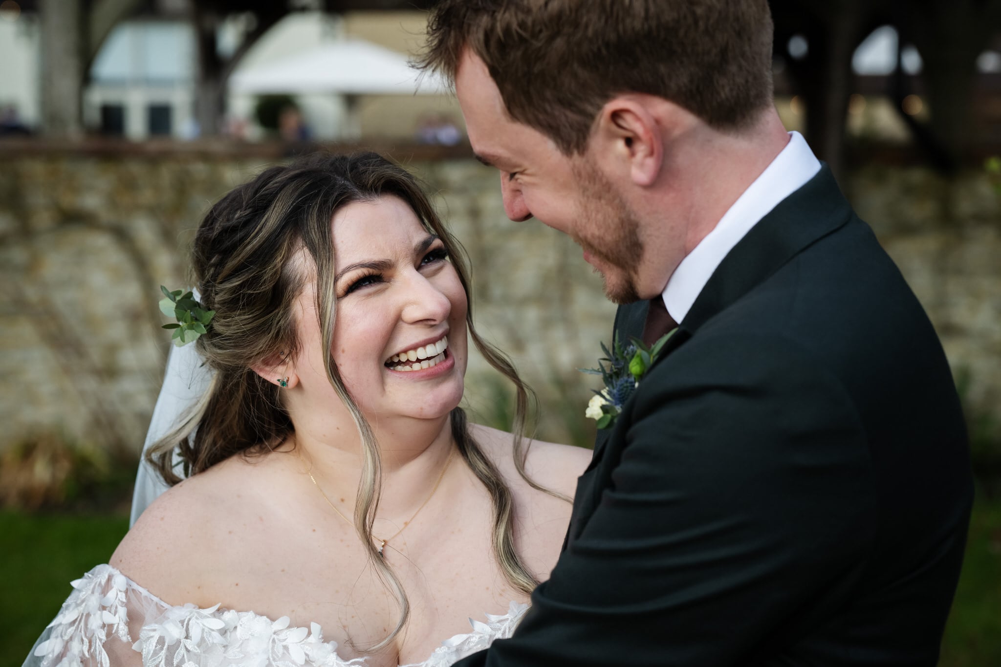 A bride and groom dressed in wedding attire smile and look at each other outdoors at Stratton Court Barn, with greenery and a stone wall in the background.