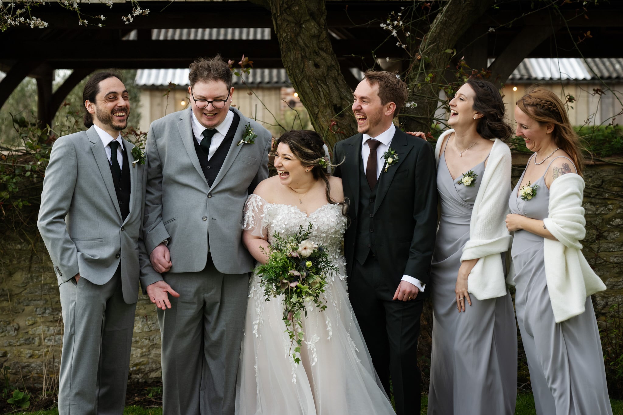 A bride and groom stand outdoors at Stratton Court Barn with three attendants, all laughing together in formal attire, surrounded by greenery and a charming stone wall.